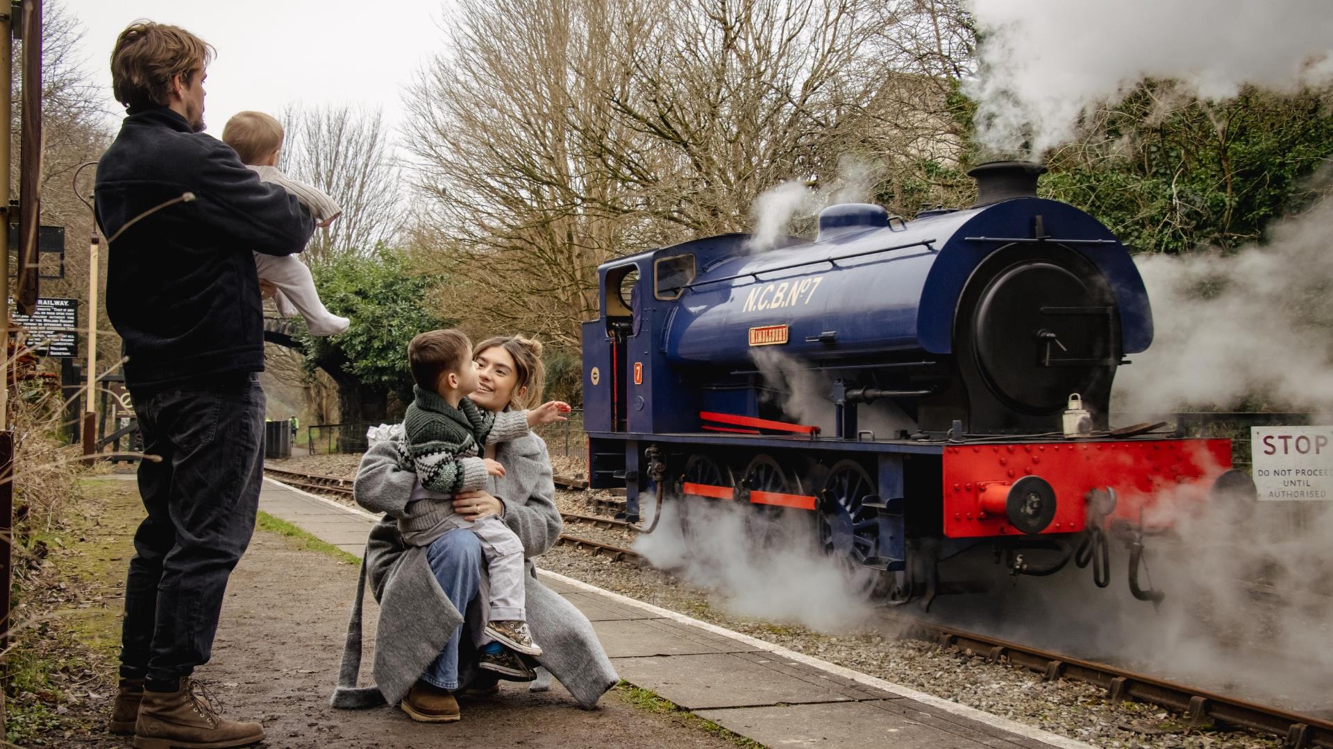 A family waiting on a railway platform with a steam train in the background.