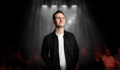 a smiling man in a black jacket stands under stage spotlights in front of a blurred audience.