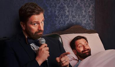 A bearded man in a tuxedo holds a microphone and points at his lookalike lying in bed