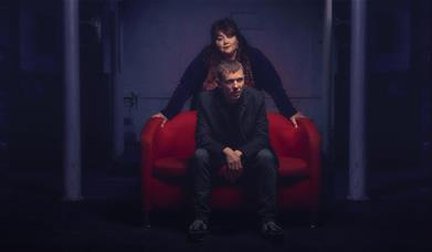 A man sits on a red sofa while a woman leans over him in a dim-lit room.