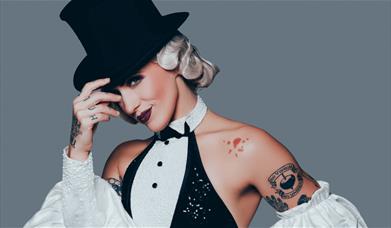 A tattooed performer in a top hat and pearl-trimmed tuxedo outfit , tips her hat with a smile.