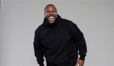 A bearded man in a black hoodie laughs heartily against a grey backgrund.