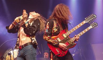 A singer with a tambourine and a guitarist with a red double-neck guitar perform under purple stage lights.