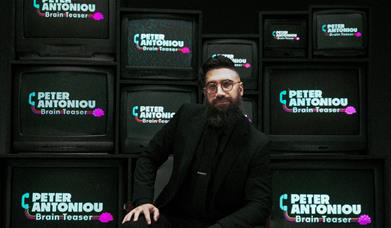 A man in round glasses and a black suit poses in front of TVs showing the Brain Teaser Logo. 