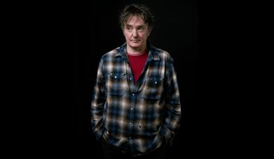 Portrait of Dylan Moran in a plaid shirt and red T-shirt standing against a black background. 