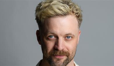 Headshot of Thomas Green with bleached blonde hair and a beard, looking at the camera against a grey background.