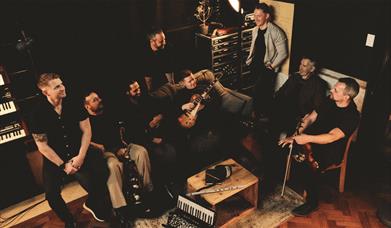 Photo of 8 band members sitting in a relaxed studio. Some of them are holding instruments including  a guitar and a violin.