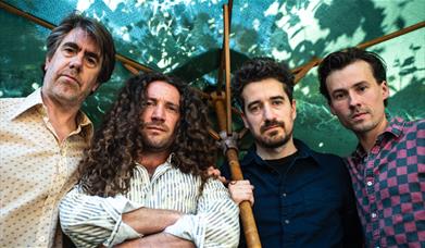 Four-piece band posed together under a large parasol against a green leafy backdrop.