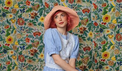 Woman in a pink sun hat posing against a colourful floral wall.