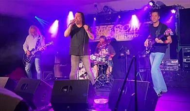 Live band performing on stage with singer, guitarist, bassist and drummer under purple lighting. 