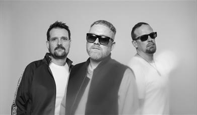 Black and white image of three men against a grey background. Two of them wearing sunglasses, all looking serious.