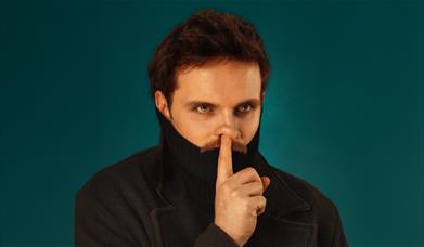 Man in dark polo neck holds a finger to his lips against a teal background. 