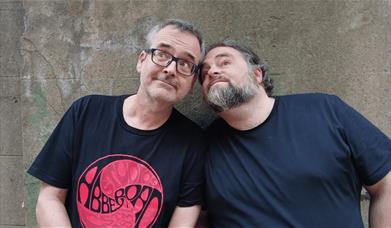 Two men in dark T-shirts leaning their heads together and pulling funny faces against a weathered stone wall.