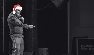 A comedian in red Santa hat points at the audience on stage in a black-and-white photo.