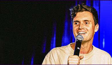 A smiling comedian holds a microphone against a blue stage curtain.
