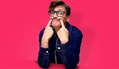 A comedian in glasses and a blue jacket pulls a silly face against a bright pink background.