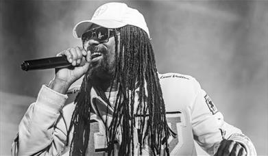 Black and white photo of a MC with long dreadlocks performing into a microphone, wearing sunglasses and a cap.