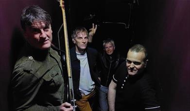 Four men posed against a dark background, one holding a guitar.