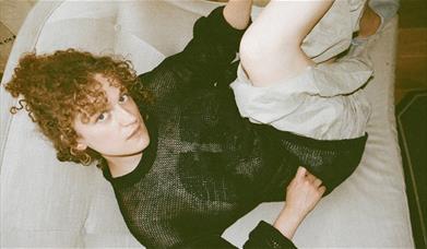 A person with curly red hair in a black mesh top lies on a white sofa, looking up at the camera.
