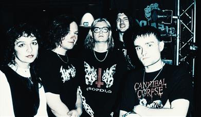Band photo of five young people backstage in dark, moody lighting, wearing black metal-band t-shirts. 