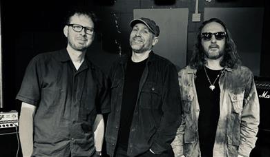 Photo of three male band members against a dark studio background. One wearing glasses, one wearing a cap and one wearing sunglasses. All smirking.