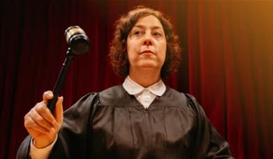 Woman dressed as a judge in robes holds up a gavel against a red curtain backdrop.