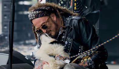 A rock musician in a studded leather jacket cuddles a fluffy dog on stage.