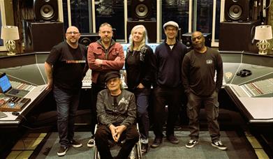 Six-piece band posing together in a recording studio, in front of a large mixing desk and studio monitors.
