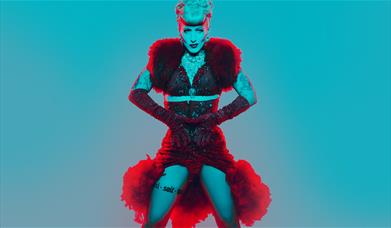A tattooed performer in a red sequinned costume with fur trim and long gloves, shot against a teal background.