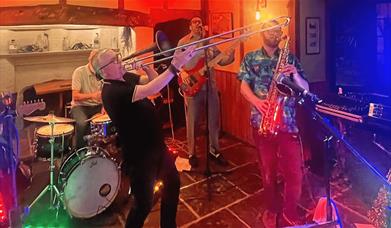 Photo of a band performing in a venue. With colourful lighting, a trumpet, saxophone, guitar and drum kit.