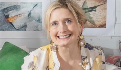 Cressida Cowell smiling with statement earrings on. Siting in front of illustrated dragon artwork on the wall.