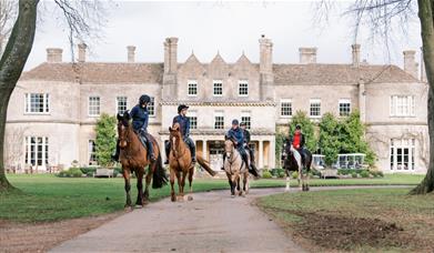 Four horses and riders coming down a path away from a large manor house hotel called Lucknam Park