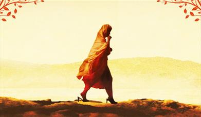 A woman in Western Asian clothing walks through a sandy terrain. 