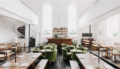 Modern restaurant interior with high ceilings, large arched windows, green cushioned chairs, wooden tables, and a central bar; a spiral staircase is v