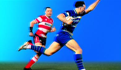 Two rugby players in blue and red kits compete for the ball in a tackle, set against a textured blue background and a grassy pitch
