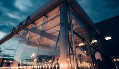 A night time image of the side of Boann distillery. The side of the distillery is all glass allowing a view into the shiny copper stills inside.