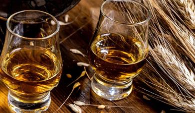 Two clear glass whisky tumblers filled with golden amber liquor, set on a rustic wooden surface with scattered grains and a bundle of wheat stalks nea