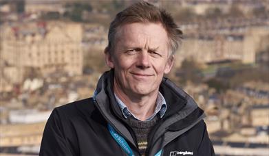 A man smiling at the camera standing above the city of Bath, wearing a Blue Badge lanyard. 