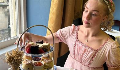 A woman in a pink regency dress reaching out towards a cake on a tiered afternoon tea platter, sat beside a window in a dining room with blue walls.