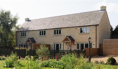 The outside of sandstone row of two cottages with a slate pitch roof and porched front doors with a brown picket fence outside and a garden full of wi