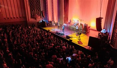 A busy crowd of combined seating and standing event attendees watch a band play live at The Forum, with red lighting and yellow spotlights.