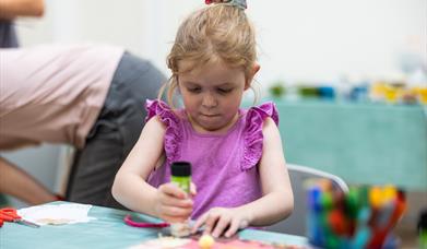 A young girl in a purple top making art with a glue stick