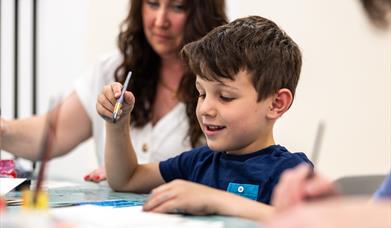 A boy making art with a paintbrush, with his mum looking on.