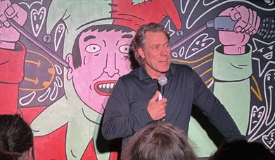 A comedian with a microphone in a black shirt, on stage against a Jester cartoon backdrop