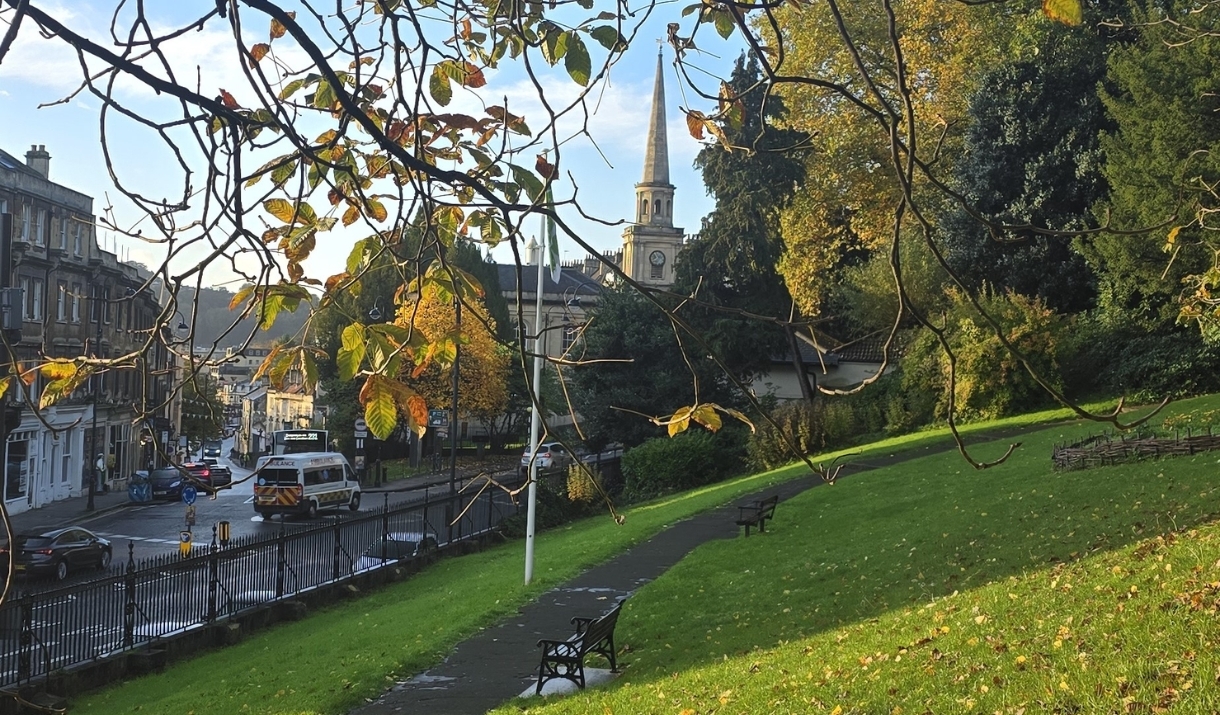 Green parkland and a footpath overlooking Bath, framed by autumn trees with a church spire in the distance.