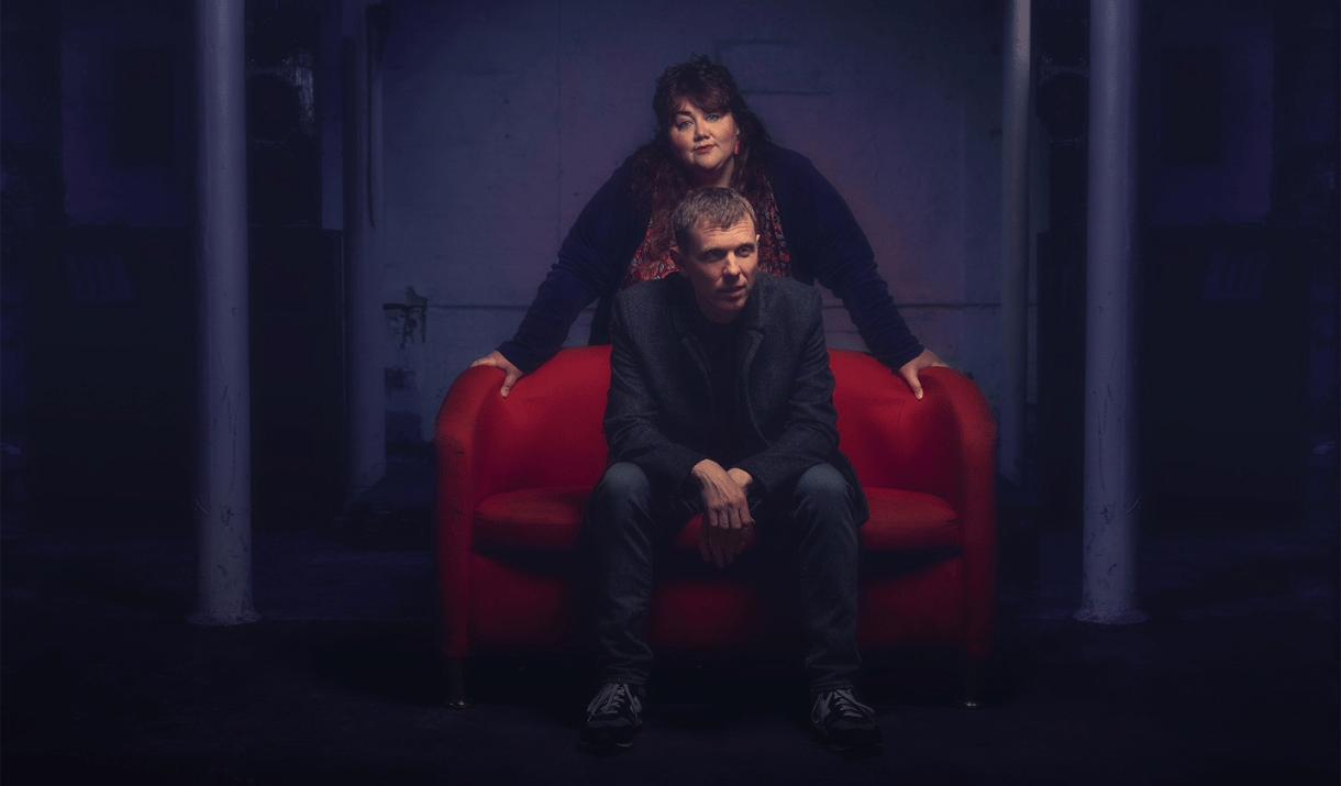 A man sits on a red sofa while a woman leans over him in a dim-lit room.