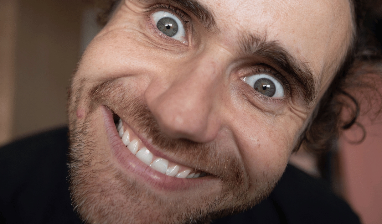 A wide-eyed man grins manically in an extreme close-up.