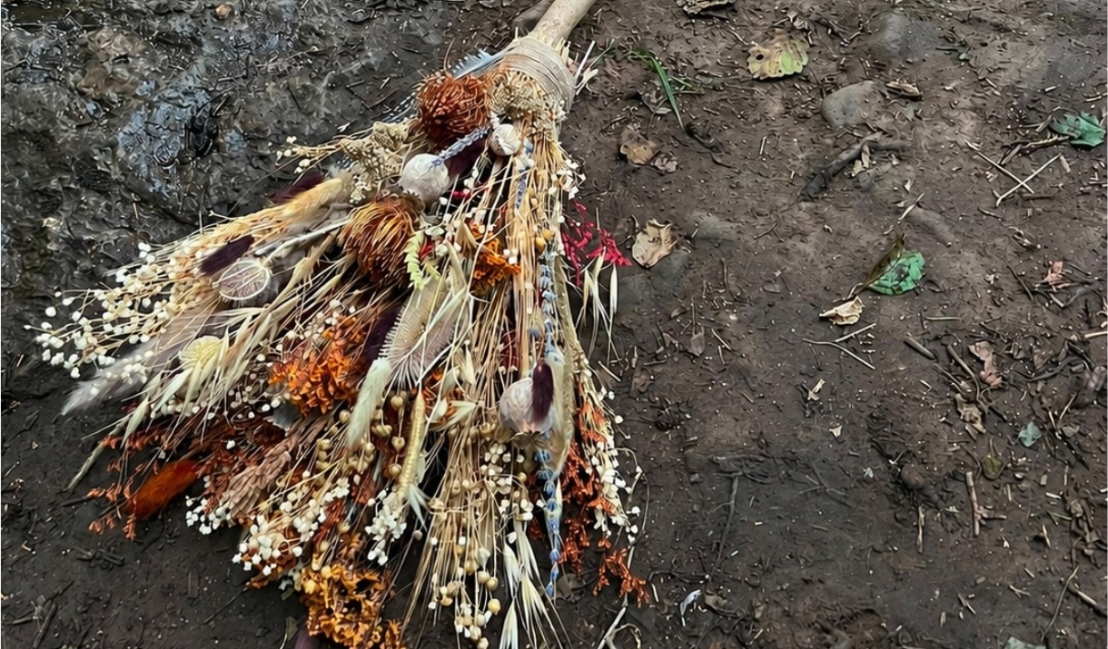 A broomstick with dried florals as the 'brush' lies on a river bank.