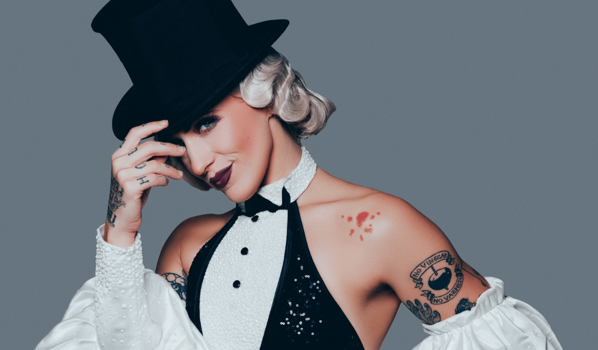A tattooed performer in a top hat and pearl-trimmed tuxedo outfit , tips her hat with a smile.