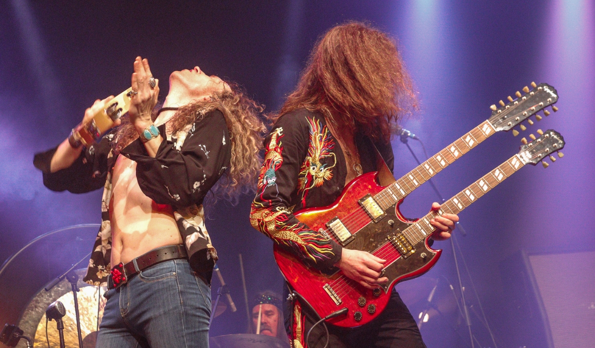 A singer with a tambourine and a guitarist with a red double-neck guitar perform under purple stage lights.